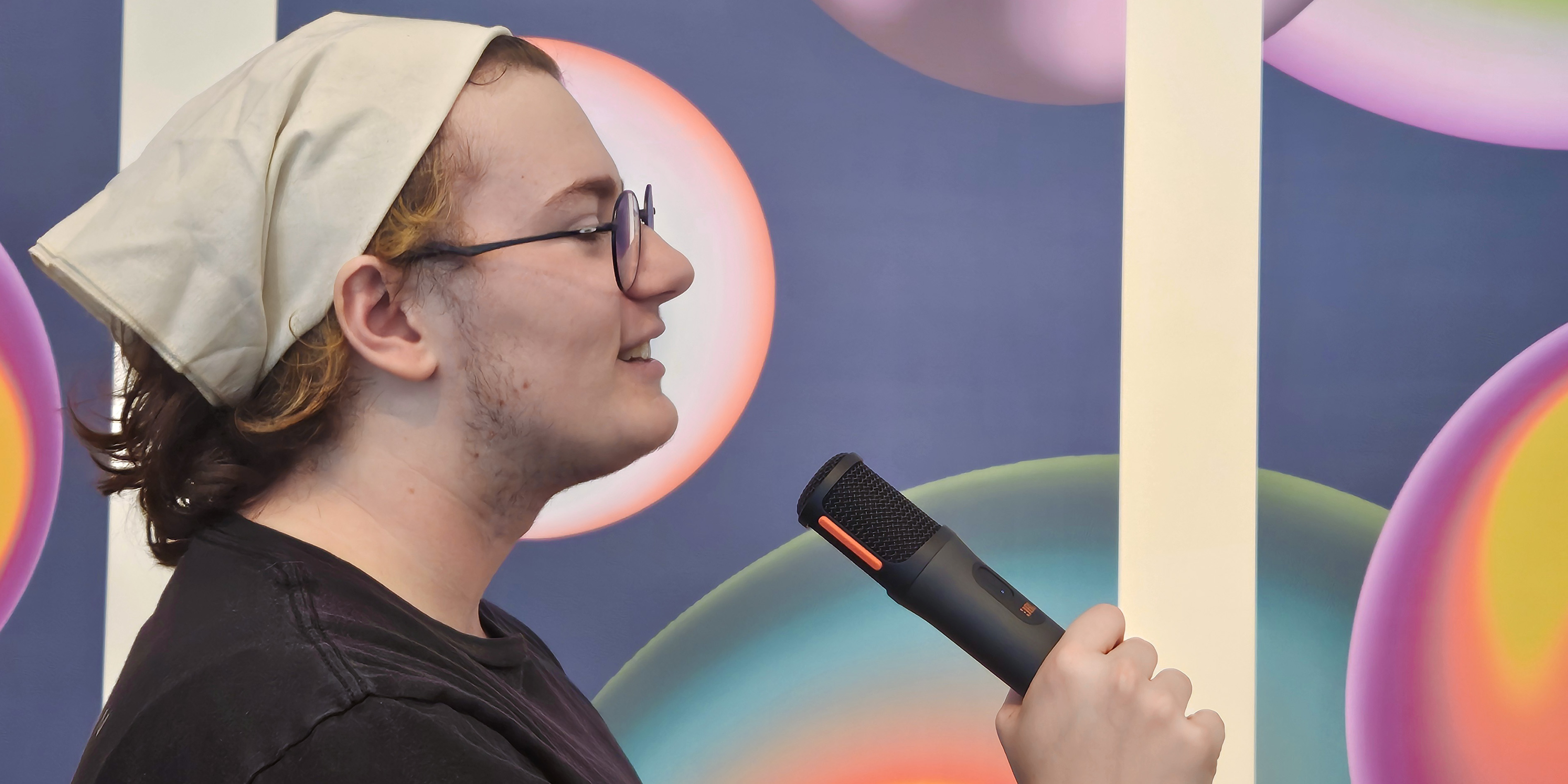 From the side facing left, a man sings holding a microphone against a blue and purple backdrop