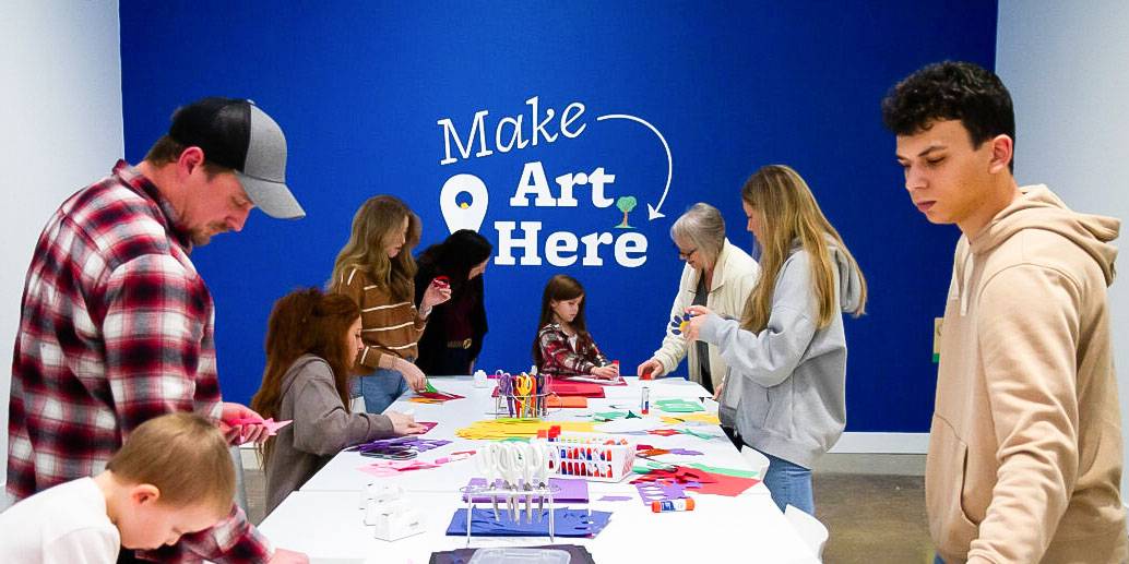 blue wall that say "make art here" pointing down to a white table covered in construction paper, scissors, and glue surrounded by people making art